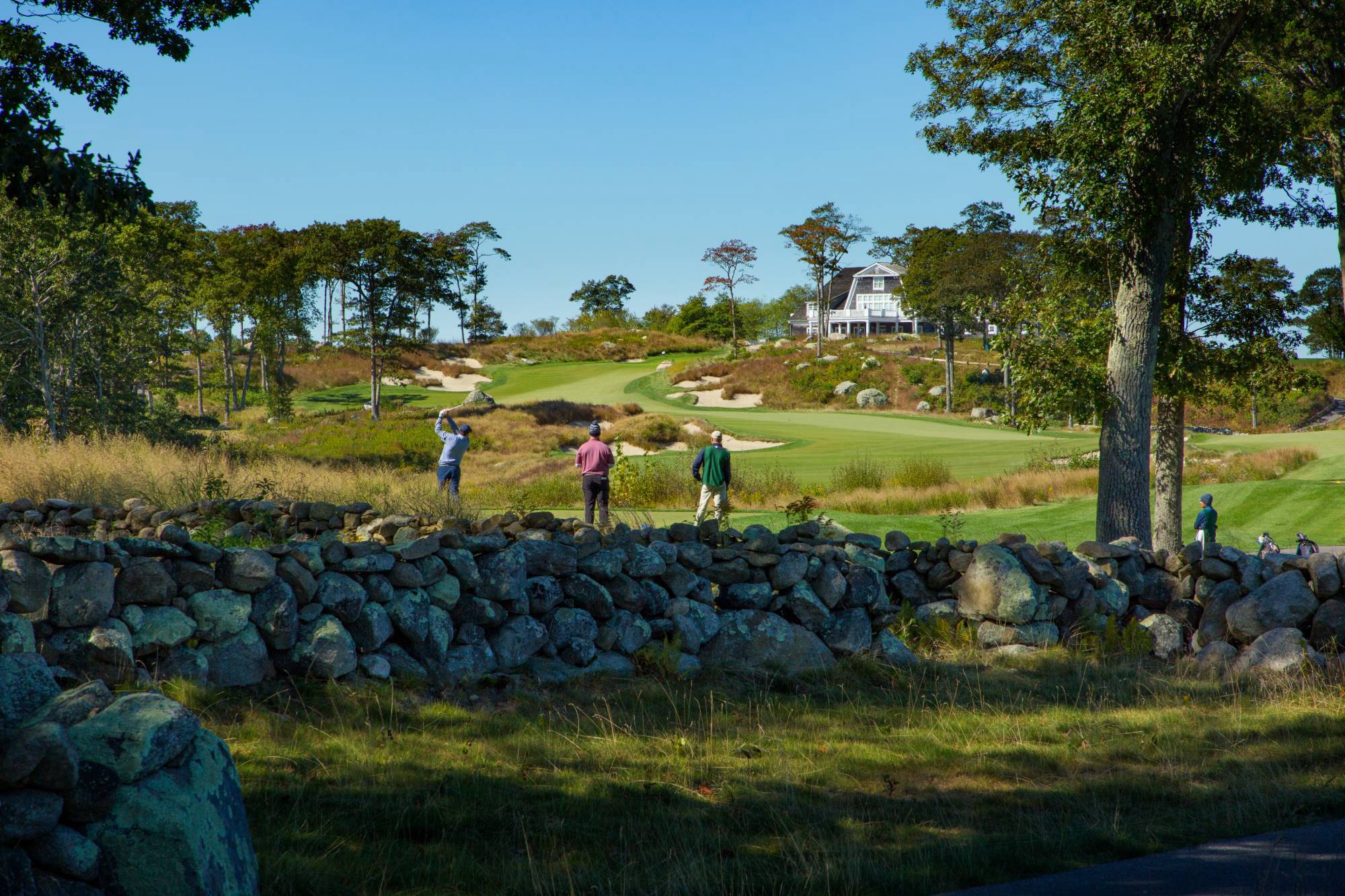 Golfers on a tee beyond a stone wall, clubhouse on the hill